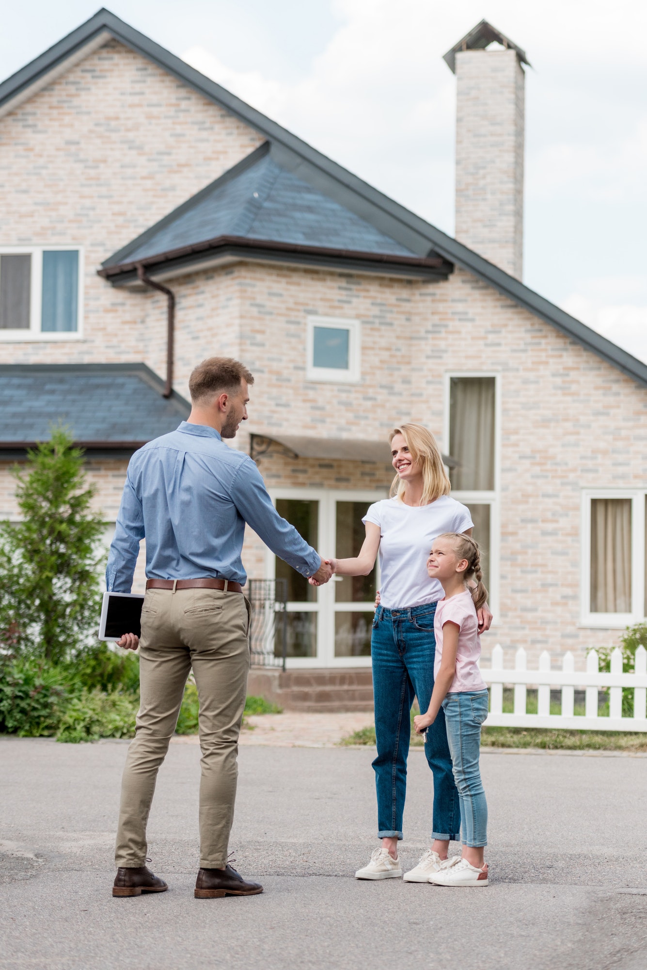 realtor with digital tablet shaking hand of woman with daughter in front of new cottage